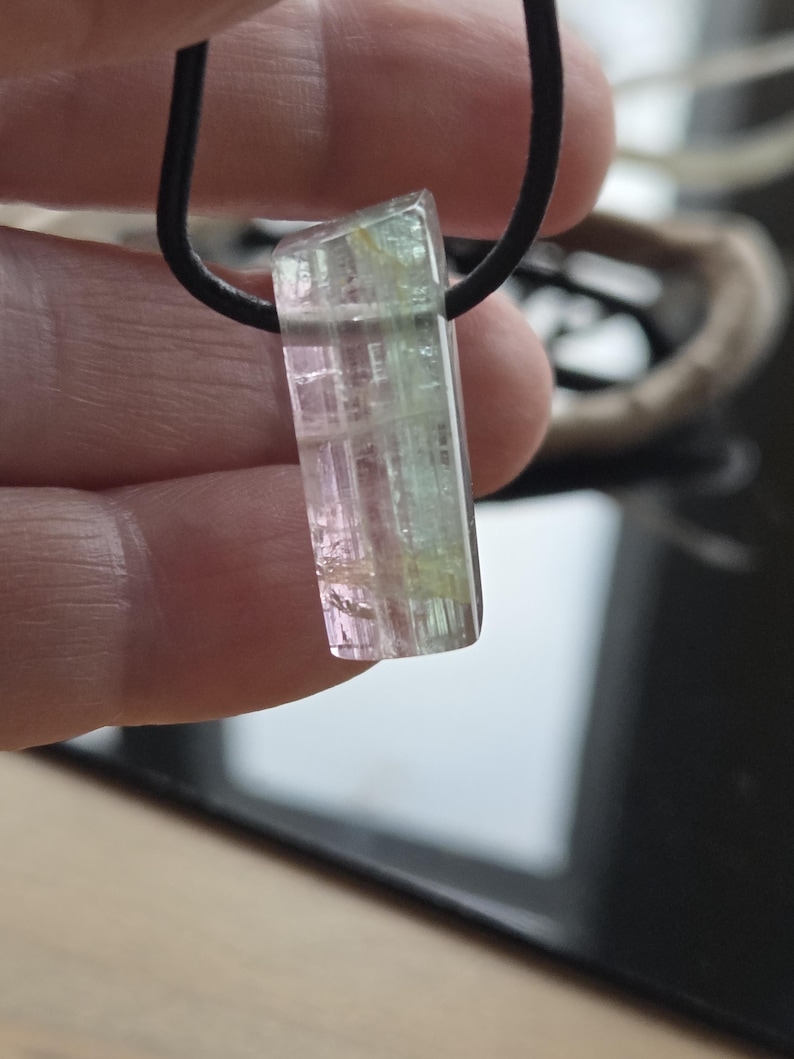 May include: A rectangular pendant necklace featuring a multi-colored tourmaline gemstone. The gemstone displays pink, green, and clear sections. It is suspended from a black cord. The pendant is approximately 1 inch long.