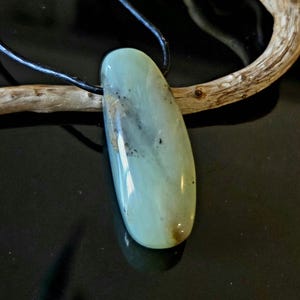 Può includere: Un pendente ovale verde chiaro con un cordino nero. La pietra presenta inclusioni marroni scure e nere. Il pendente è esposto su una superficie nera con un pezzo di legno alla deriva.