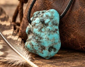 May include: A raw, irregular turquoise stone pendant featuring vibrant blue-green hues with dark grey matrix inclusions, suspended from a simple black cord. The stone has a natural, unpolished texture with visible pits and uneven edges.