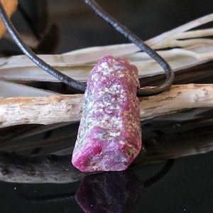 Op de afbeelding: Een ruwe robijn hanger aan een zwart leren koord. De robijn heeft een diep roze-rode kleur met een ruw, getextureerd oppervlak. De hanger hangt tegen een donkere achtergrond met een houten tak.