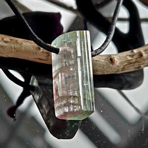 May include: A rectangular tourmaline pendant with a black cord. The gemstone displays a gradient of colors, including green, pink, and clear sections. The pendant is suspended against a reflective surface, with a wooden branch and a dark purple leaf in the background.
