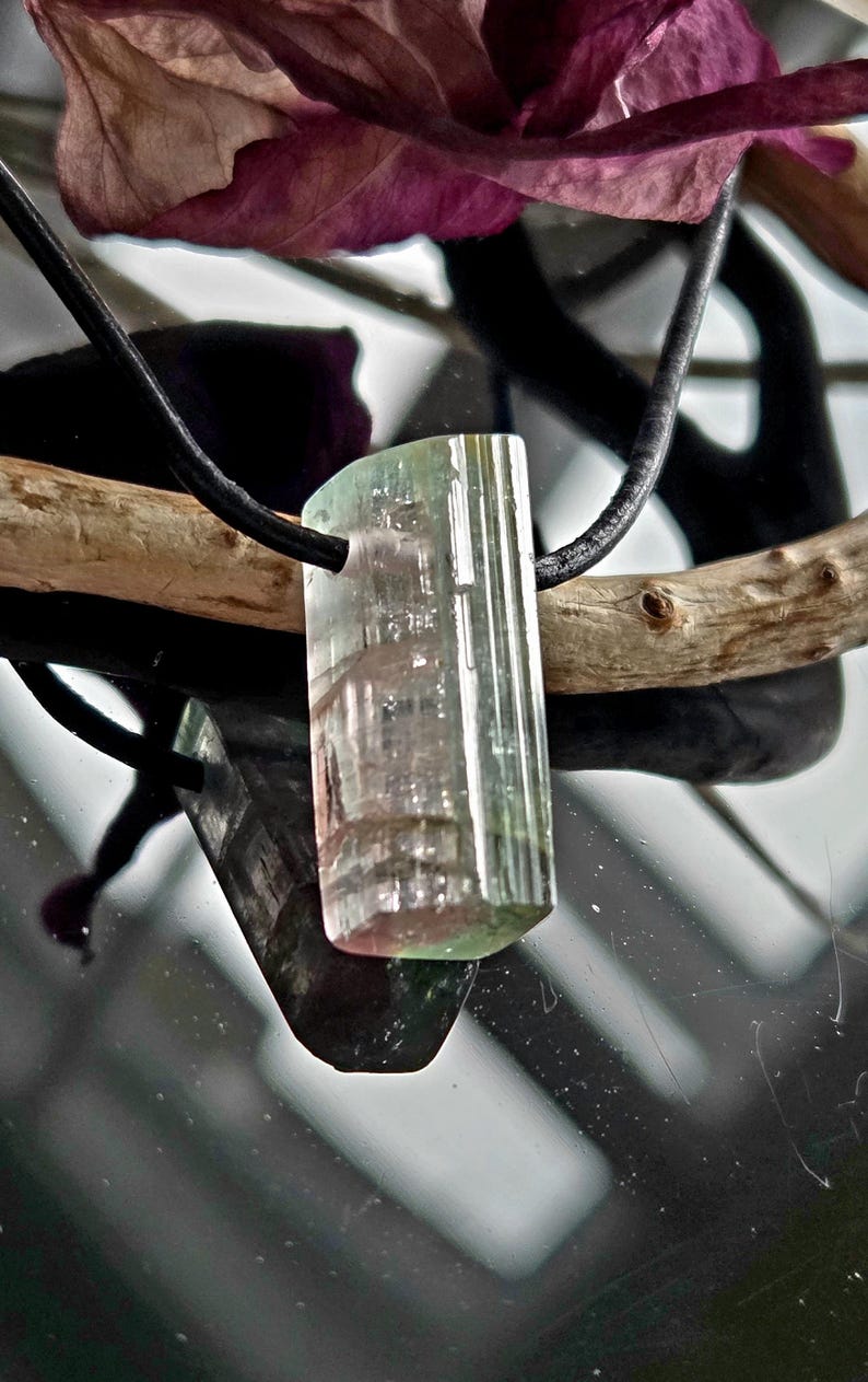 May include: A rectangular tourmaline pendant with a black cord. The gemstone displays a gradient of colors, including green, pink, and clear sections. The pendant is suspended against a reflective surface, with a wooden branch and a dark purple leaf in the background.