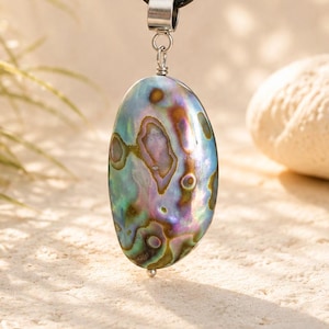 Peut inclure: Un pendentif ovale en coquille d'ormeau présentant des tourbillons irisés de bleu, vert, violet et marron. Le pendentif est suspendu à un cordon noir par une bélière en métal argenté et comporte une petite perle argentée sur le bord inférieur.