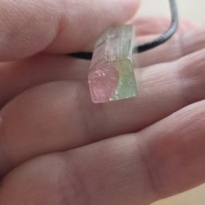 May include: A watermelon tourmaline pendant with a black cord. The crystal is a rectangular shape with pink, green, and clear sections. The pendant is held in a hand.