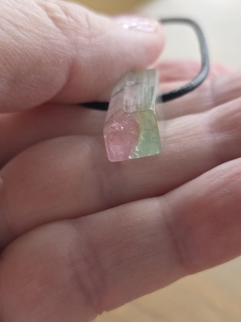 May include: A watermelon tourmaline pendant with a black cord. The crystal is a rectangular shape with pink, green, and clear sections. The pendant is held in a hand.