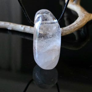 Puede incluir: Un colgante de cuarzo transparente con un cordón negro. La piedra pulida, de forma ovalada, es translúcida, con inclusiones internas en forma de nube. El colgante cuelga sobre un fondo oscuro, con una rama visible.