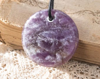 May include: A circular purple pendant with a polished, textured surface featuring natural mineral inclusions. The pendant has a silver-toned metal grommet at the top and is suspended from a smooth black cord, creating a simple necklace design.