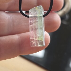May include: A clear, rectangular crystal pendant with pink and green hues, suspended from a black cord. The crystal has a textured appearance and is held up by a hand. The pendant is a unique piece of jewelry.