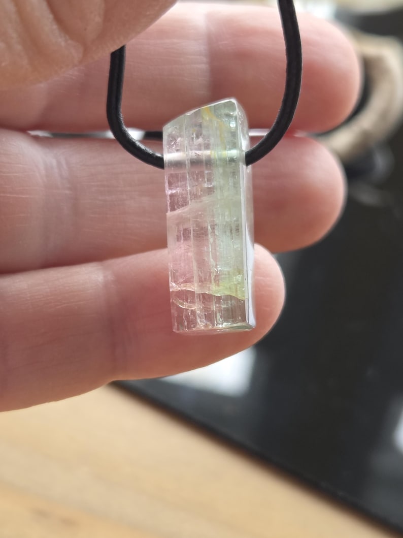 May include: A clear, rectangular crystal pendant with pink and green hues, suspended from a black cord. The crystal has a textured appearance and is held up by a hand. The pendant is a unique piece of jewelry.