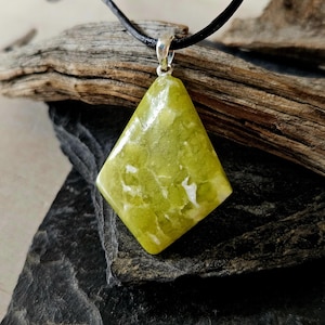 Peut inclure: Un collier pendentif en forme de losange avec une pierre verte vive et des veines blanches. Le pendentif est suspendu à un cordon en cuir noir et à une bélière argentée. Le collier est présenté sur une surface sombre et texturée.