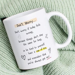 May include: White ceramic mug with a curved handle. The mug features the text "Don't Worry..." followed by an inspirational message and heart illustrations. The mug is set against a light green knitted background.
