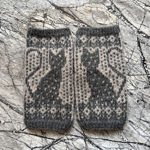 May include: A pair of charcoal gray and light beige knit fingerless gloves featuring a repeating fair isle pattern of a sitting cat silhouette. The cuffs and edges are finished with a ribbed knit texture, showcasing a detailed two-tone design.