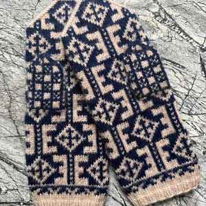 Puede incluir: Un par de manoplas tejidas con un patrón geométrico de dos tonos en azul marino oscuro y beige. El diseño consiste en motivos repetitivos de diamantes y cruces en la mano y el pulgar, terminados con un puño acanalado en el borde inferior.