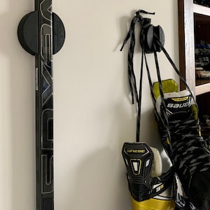 A close up of two TwigRigs mounted to the wall in a bedroom. One of them is holding a Bauer Nexus hockey stick and the other has a pair of Bauer Supreme skates hanging from it.