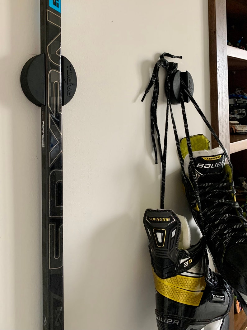 A close up of two TwigRigs mounted to the wall in a bedroom. One of them is holding a Bauer Nexus hockey stick and the other has a pair of Bauer Supreme skates hanging from it.