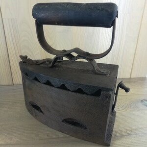 May include: A vintage cast iron flat iron with a wooden handle. The iron has a hinged door and a small lever on the side. The iron is rusty and shows signs of wear.