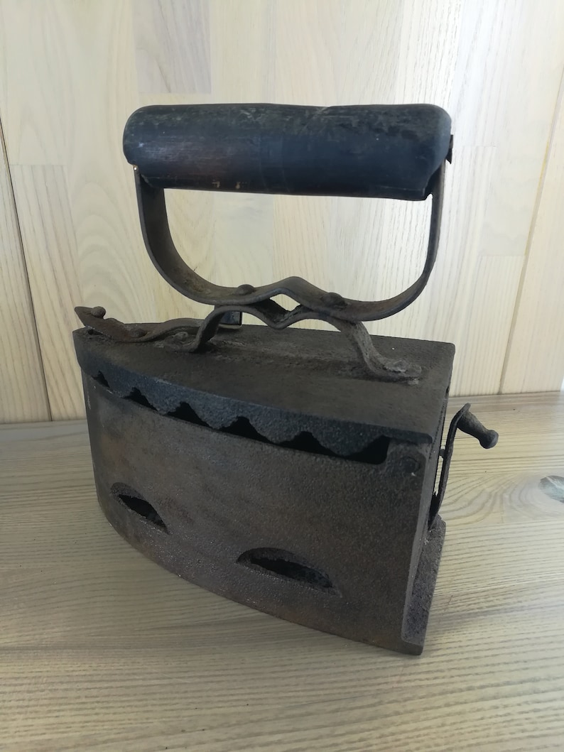 May include: A vintage cast iron flat iron with a wooden handle. The iron has a hinged door and a small lever on the side. The iron is rusty and shows signs of wear.