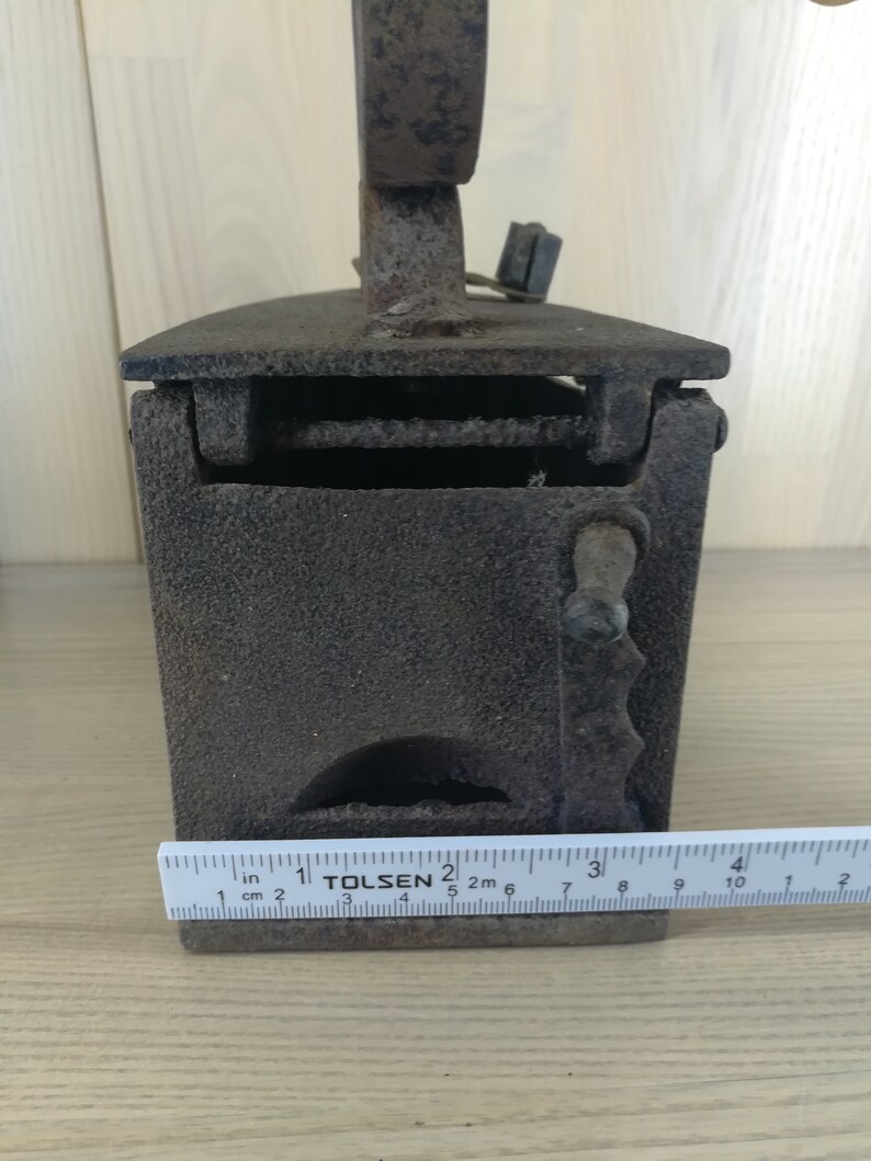 May include: A vintage cast iron charcoal iron with a hinged top and a handle. The iron is approximately 10 inches long and has a ruler for measuring in inches and centimeters.