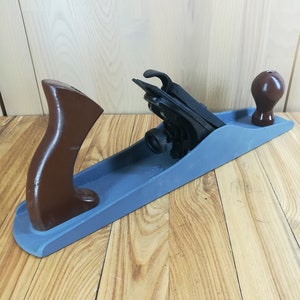 Puede incluir: Un cepillo de mano de madera azul con un mango marrón y una cuchilla de metal negro. El cepillo está sobre una superficie de madera.