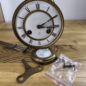 May include: A round brass clock face with a white dial, black Roman numerals, and two black hands. It features a pendulum with a circular white insert, a brass winding key, and a small plastic bag containing three black metal screws.