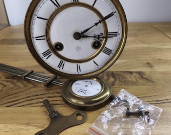 Op de afbeelding: Een ronde messing klokplaat met een witte wijzerplaat, zwarte Romeinse cijfers en twee zwarte wijzers. Het bevat een slinger met een ronde witte inzet, een messing opwindsleutel en een plastic zakje met drie zwarte metalen schroeven.
