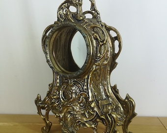 Op de afbeelding: Een gouden messing klokkenstandaard met ingewikkelde, sierlijke details en een rond, gespiegeld gezicht. De standaard heeft drie poten en een decoratieve bovenkant.