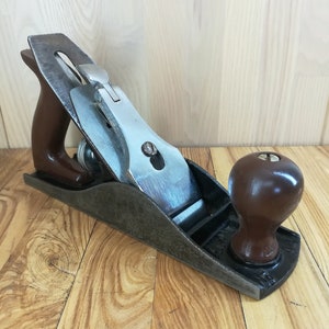 May include: A classic hand plane, featuring a dark brown wooden handle and knob. The metal blade and body show signs of wear. This woodworking tool is designed for smoothing surfaces, with a blade and body made of metal.