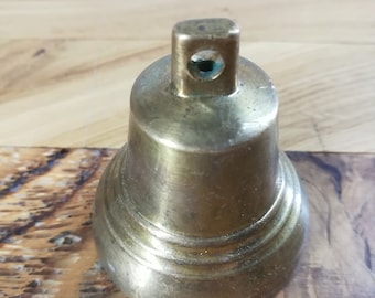 Pode incluir: Um pequeno sino de latão com uma base arredondada e um topo quadrado. O sino tem uma superfície lisa e polida e um pequeno furo no topo.