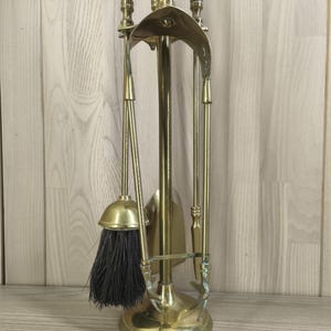 May include: A brass fireplace tool set featuring a central stand with a circular base, holding a brush with dark bristles, a shovel, and a poker. Each tool has a decorative handle with a polished metallic finish and a curved top support.