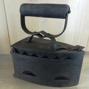May include: A vintage cast iron flat iron with a wooden handle. The iron has a scalloped edge and several air vents.