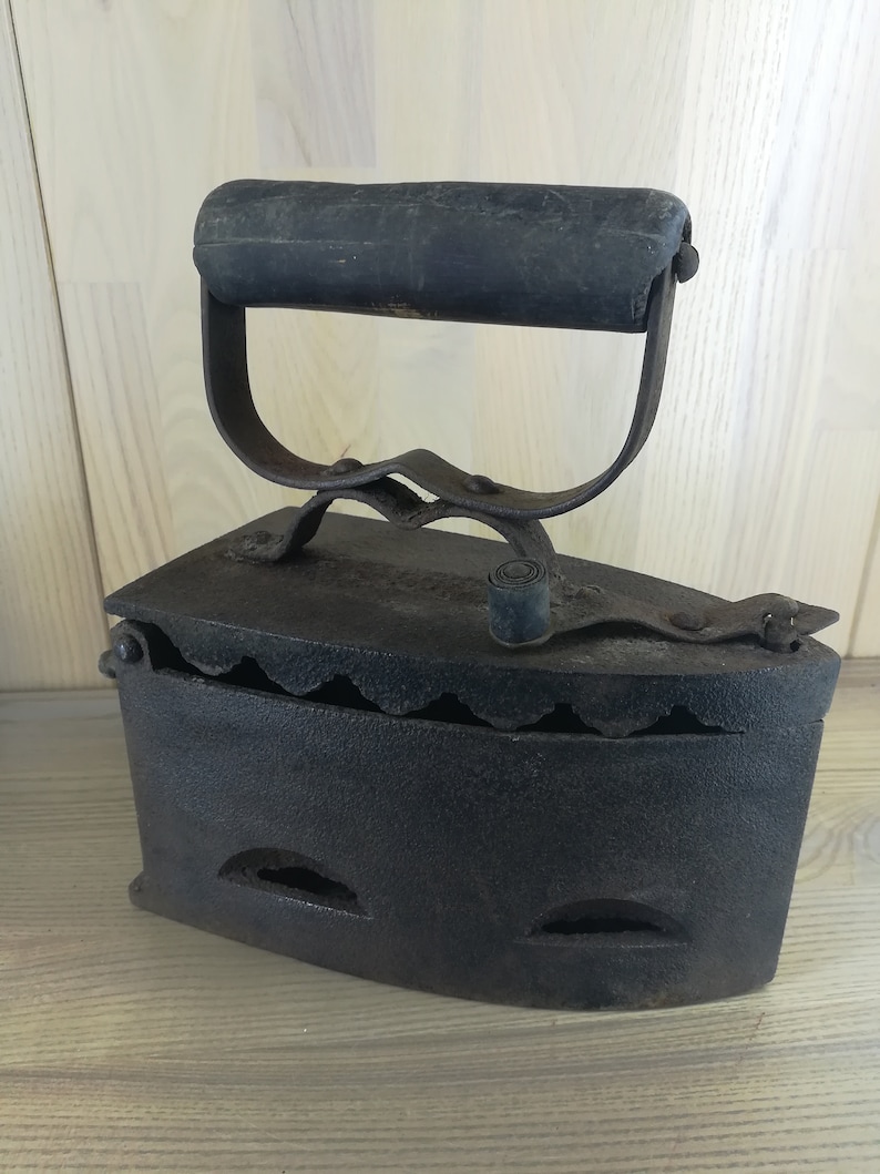 May include: A vintage cast iron flat iron with a wooden handle. The iron has a scalloped edge and several air vents.