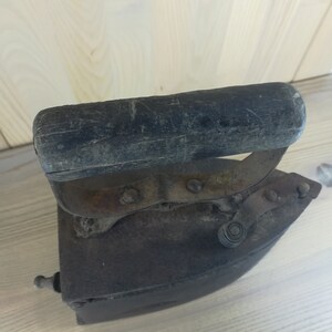 May include: An antique cast iron flat iron with a wooden handle. The iron is rusty and has a worn finish. The handle is dark brown and has a worn finish.