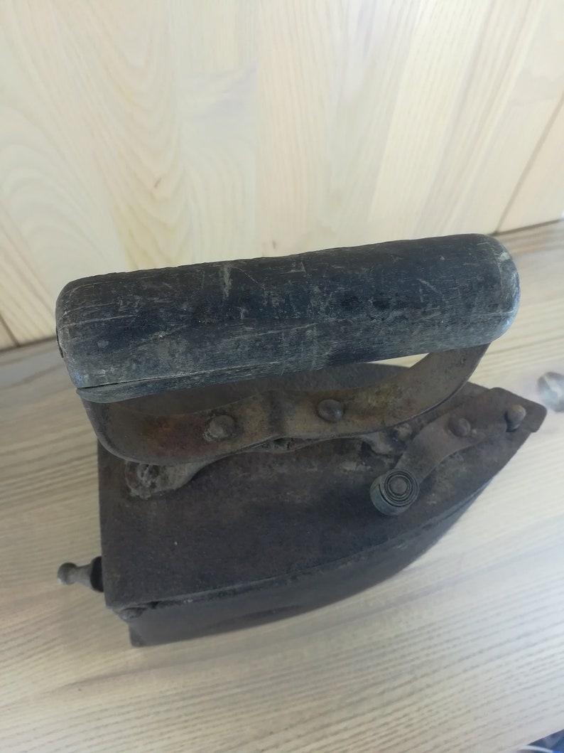 May include: An antique cast iron flat iron with a wooden handle. The iron is rusty and has a worn finish. The handle is dark brown and has a worn finish.