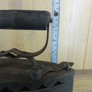 May include: A vintage cast iron flat iron with a wooden handle and a metal latch. The iron is approximately 8 inches tall and has a black finish.
