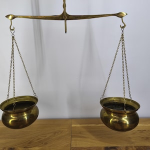 Könnte beinhalten: Eine antike Messing-Balkenwaage mit einem zentralen horizontalen Balken, einem Zeiger und zwei hängenden Schalen, die an dünnen Metallketten befestigt sind. Die gesamte Konstruktion besteht aus poliertem Messing mit einem warmen Goldton.