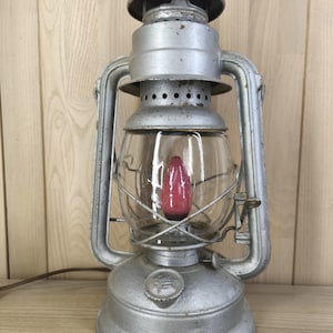 May include: A silver-colored metal lantern with a clear glass globe and a pink flame-shaped light bulb inside. The lantern features a top handle, a circular base, and a brown electrical cord extending from the side of the base unit.