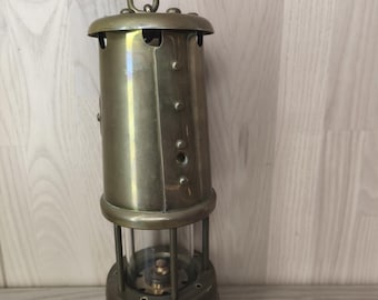 Op de afbeelding: Een koperen mijnwerkerslamp met een cilindervormig lichaam, een middengedeelte van helder glas, een massieve metalen voet en een gebogen ophanghaak aan de bovenkant. Het metalen oppervlak heeft een gepolijste afwerking met zichtbare klinknagels.
