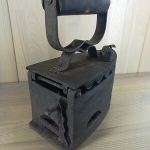 May include: A vintage cast iron charcoal iron with a wooden handle. The iron has a hinged door and a grate for burning charcoal.