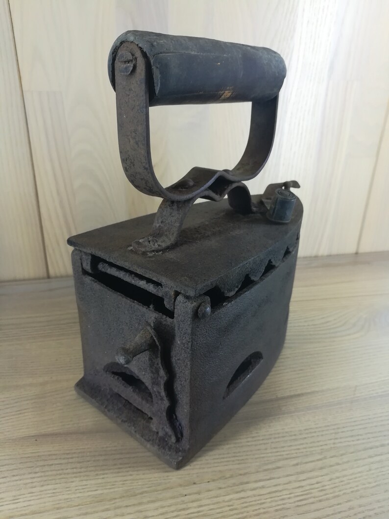 May include: A vintage cast iron charcoal iron with a wooden handle. The iron has a hinged door and a grate for burning charcoal.