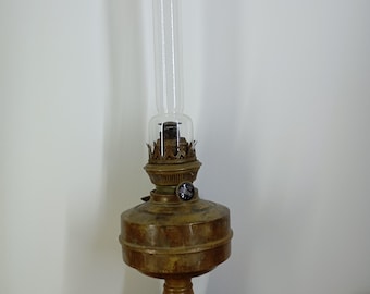 Op de afbeelding: Een antieke olielamp met een heldere glazen schoorsteen en een bronzen metalen voet. De lamp heeft een rond reservoir, een decoratieve bovenkant en een cirkelvormige voet. De lamp is ontworpen om op een vlakke ondergrond te staan.