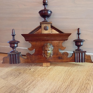 Könnte beinhalten: Dunkelbraunes dekoratives Wanduhrstück aus Holz mit aufwendigen Schnitzereien. Die Uhr hat eine zentrale Gesichtsschnitzerei, zwei seitliche Zierknöpfe und einen oberen Zierknopf. Das Holz hat ein sattes, gealtertes Aussehen, das auf einen Vintage- oder antiken Stil hindeutet.