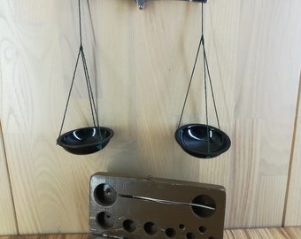 Puede incluir: Un conjunto vintage de balanzas con una caja de madera que contiene un juego de pesas. Las balanzas tienen cuencos de metal negro y están suspendidas de una barra de metal. La caja de madera está abierta y revela una variedad de pesas de diferentes tamaños.