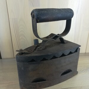 May include: A vintage cast iron coal-burning flat iron with a wooden handle. The iron has a scalloped edge and several holes on the bottom.