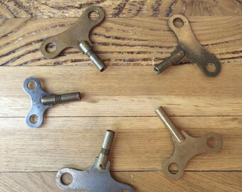 Op de afbeelding: Een verzameling van vijf metalen opwindsleutels voor klokken, gerangschikt in een cirkelvormig patroon. De sleutels hebben verschillende afwerkingen, waaronder messing- en zilverkleuren, elk met een T-vormige handgreep en een centrale cilindrische schacht.