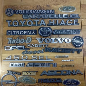 Può includere: Una collezione di emblemi e distintivi di auto d'epoca, tra cui Ford, Volkswagen, Toyota, Citroën, Volvo, Opel e Saab. Gli emblemi metallici sono disposti su una superficie di legno, mostrando vari design.