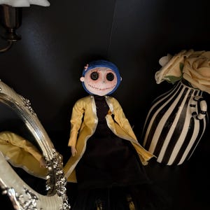 K&ouml;nnte beinhalten: Eine handgefertigte Puppe mit Knopfaugen, blauen Haaren und einem gelben Mantel &uuml;ber einem schwarzen Kleid. Die Puppe ist mit einem dekorativen Spiegel und einer schwarz-wei&szlig; gestreiften Vase mit einer cremefarbenen Blume drapiert.