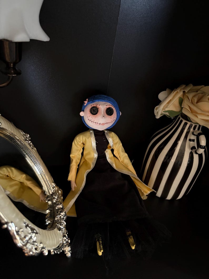 K&ouml;nnte beinhalten: Eine handgefertigte Puppe mit Knopfaugen, blauen Haaren und einem gelben Mantel &uuml;ber einem schwarzen Kleid. Die Puppe ist mit einem dekorativen Spiegel und einer schwarz-wei&szlig; gestreiften Vase mit einer cremefarbenen Blume drapiert.