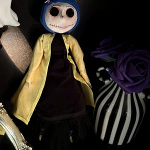K&ouml;nnte beinhalten: Handgefertigte Puppe mit bemaltem Gesicht, Knopfaugen und blauen Garnhaaren. Die Puppe tr&auml;gt ein schwarzes Kleid mit einem gelben Mantel. Eine schwarz-wei&szlig; gestreifte Vase enth&auml;lt lila Rosen. Die Puppe ist eine Figur aus dem Film Coraline.