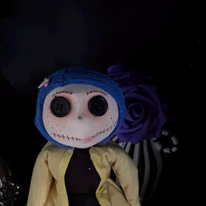 K&ouml;nnte beinhalten: Handgefertigte Puppe mit blassem Gesicht, Knopfaugen und gen&auml;htem Mund. Die Puppe hat blaue Garnhaare, einen gelben Mantel und ein schwarzes Kleid. Eine lila Rose und ein gestreiftes Band befinden sich im Hintergrund.