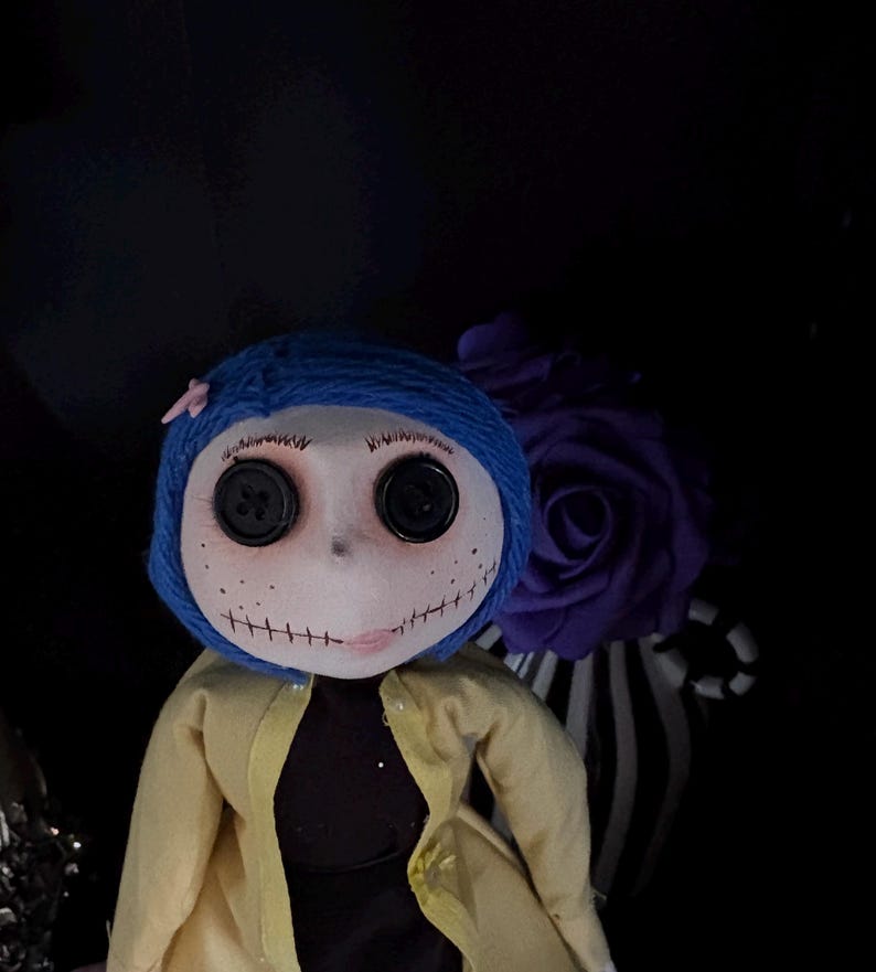K&ouml;nnte beinhalten: Handgefertigte Puppe mit blassem Gesicht, Knopfaugen und gen&auml;htem Mund. Die Puppe hat blaue Garnhaare, einen gelben Mantel und ein schwarzes Kleid. Eine lila Rose und ein gestreiftes Band befinden sich im Hintergrund.
